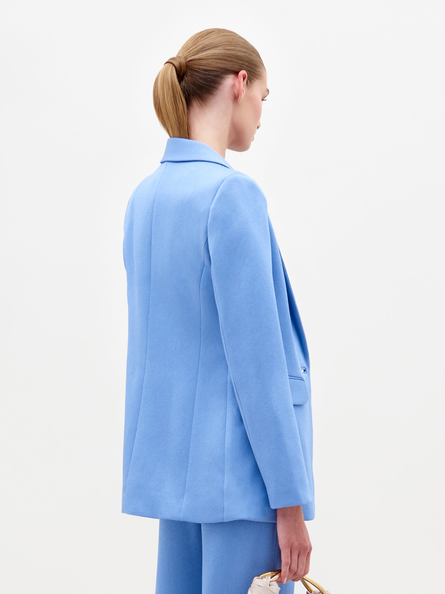 A person with blond hair in a ponytail models SVT DESIGNS' light blue Rebeca Jacket with matching pants, featuring a single-button closure. Holding a purse, they stand against a plain white background, slightly turned from the camera.