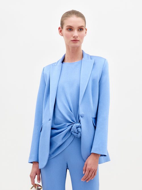 The woman wears the Rebeca Jacket from SVT DESIGNS, crafted from luxurious Italian textiles in light blue, featuring a single-button front closure and a knotted shirt. With her hair pulled back, she holds eyeglasses in one hand against a plain, light-colored background.