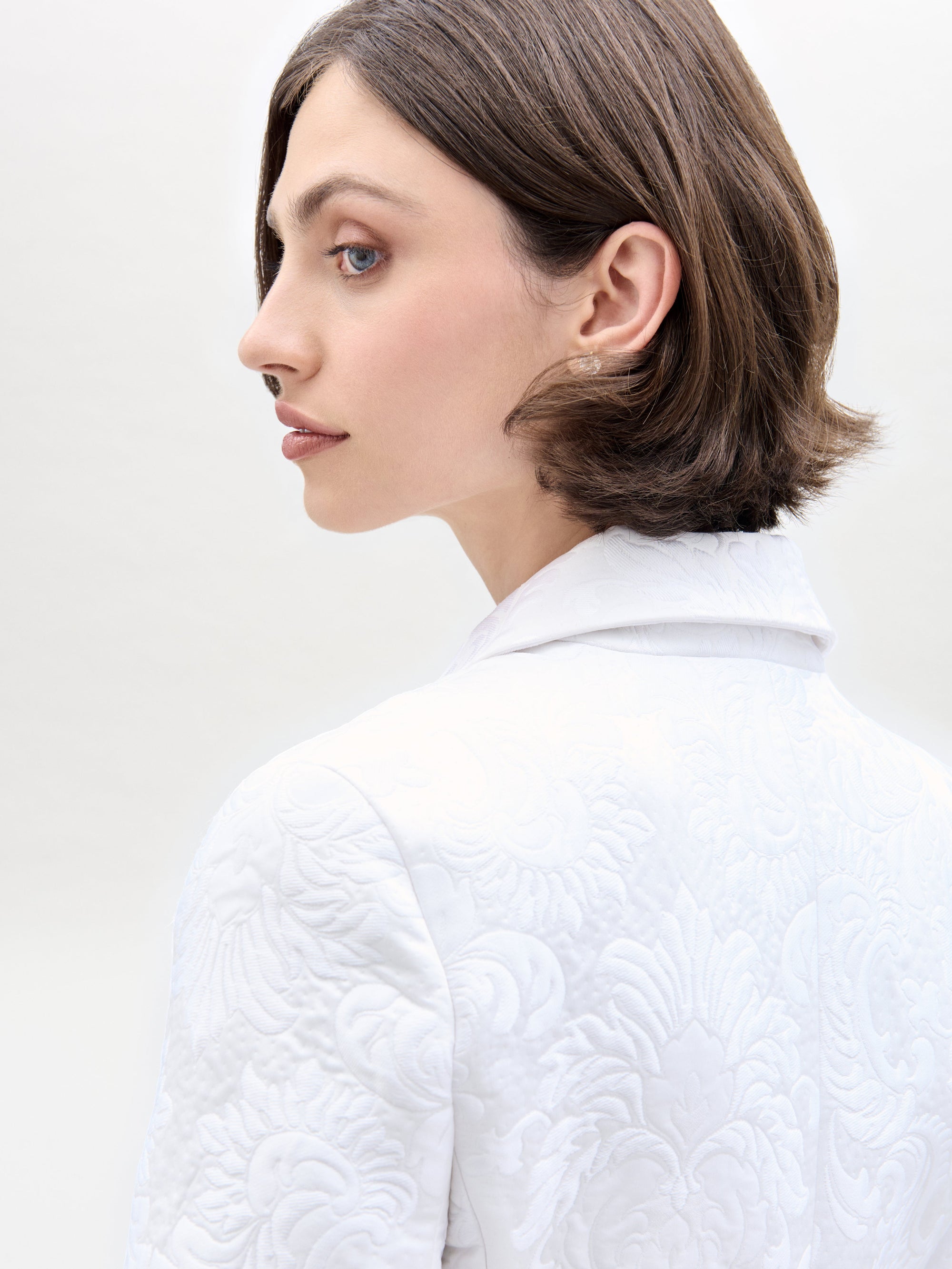 A person with short brown hair wears a white Rebeca Jacket from SVT DESIGNS, featuring a textured, intricate pattern. They show an elegant earring while facing to the side against a plain, light-colored background.