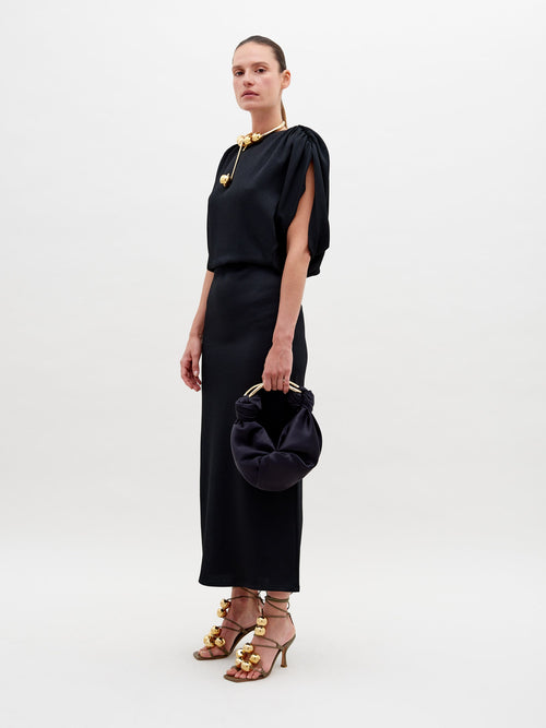 Against a plain background, a person stands elegantly in the Reese Dress Black with gold accessories, holding a matching black handbag. They also sport high-heeled sandals adorned with gold embellishments. Pre-order now to ensure your look is ready by the ship-by date of November 15th, 2024.