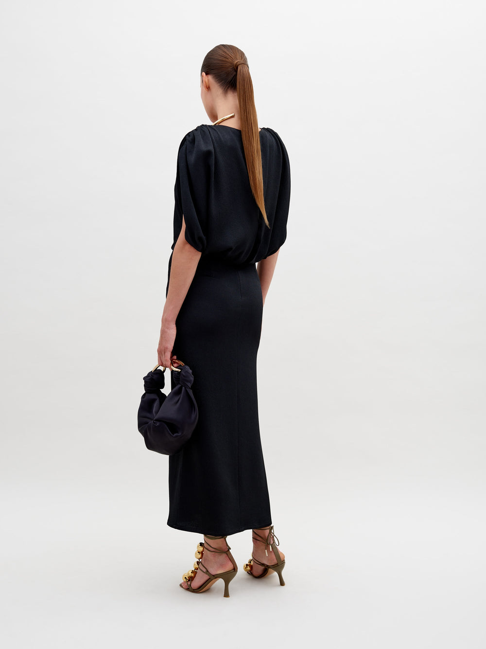 A woman with long straight hair wears the Reese Dress, a black hammered satin midi, and heeled sandals. She faces away, holding a small black handbag in her left hand against a plain white background.
