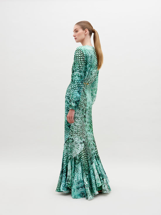 A person stands wearing the Remy Dress Emerald Animal Print, a green long-sleeve, full-length dress with a pattern resembling scales, featuring a V-neckline and a belt with ornamental details. Available for pre-order now, this stunning piece will ship by date November 15th, 2024. The background is plain white.