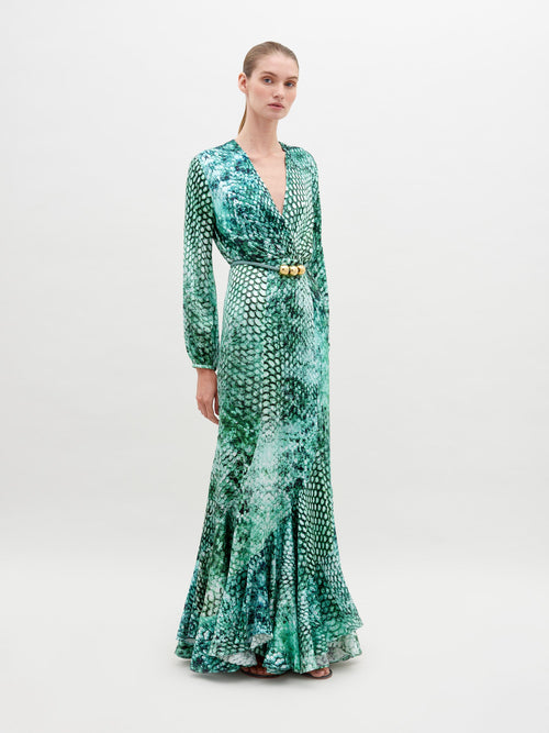 A person stands wearing the Remy Dress Emerald Animal Print, a green long-sleeve, full-length dress with a pattern resembling scales, featuring a V-neckline and a belt with ornamental details. Available for pre-order now, this stunning piece will ship by date November 15th, 2024. The background is plain white.