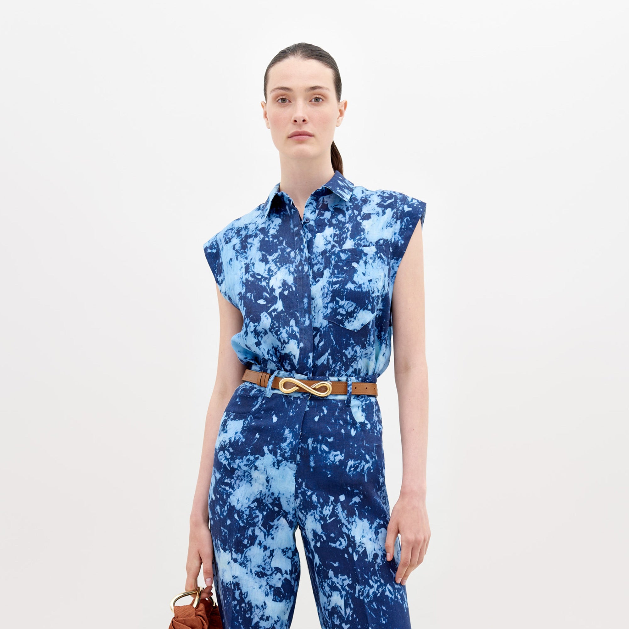 With long dark hair tied back, a person wears the Rina Blouse from SVT DESIGNS, styled with a sleeveless blue tie-dye jumpsuit made from Italian textiles and a brown infinity buckle belt. They hold a small brown bag against a plain white background.