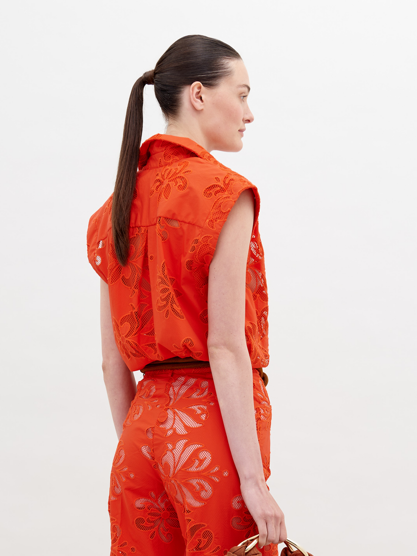 A person stands wearing a vibrant orange sleeveless top and matching pants with floral patterns, accessorized with a brown belt and holding a brown handbag. Pre-order the Rina Blouse Rouge Embroidery now to ensure it ships by the date of November 15th, 2024.