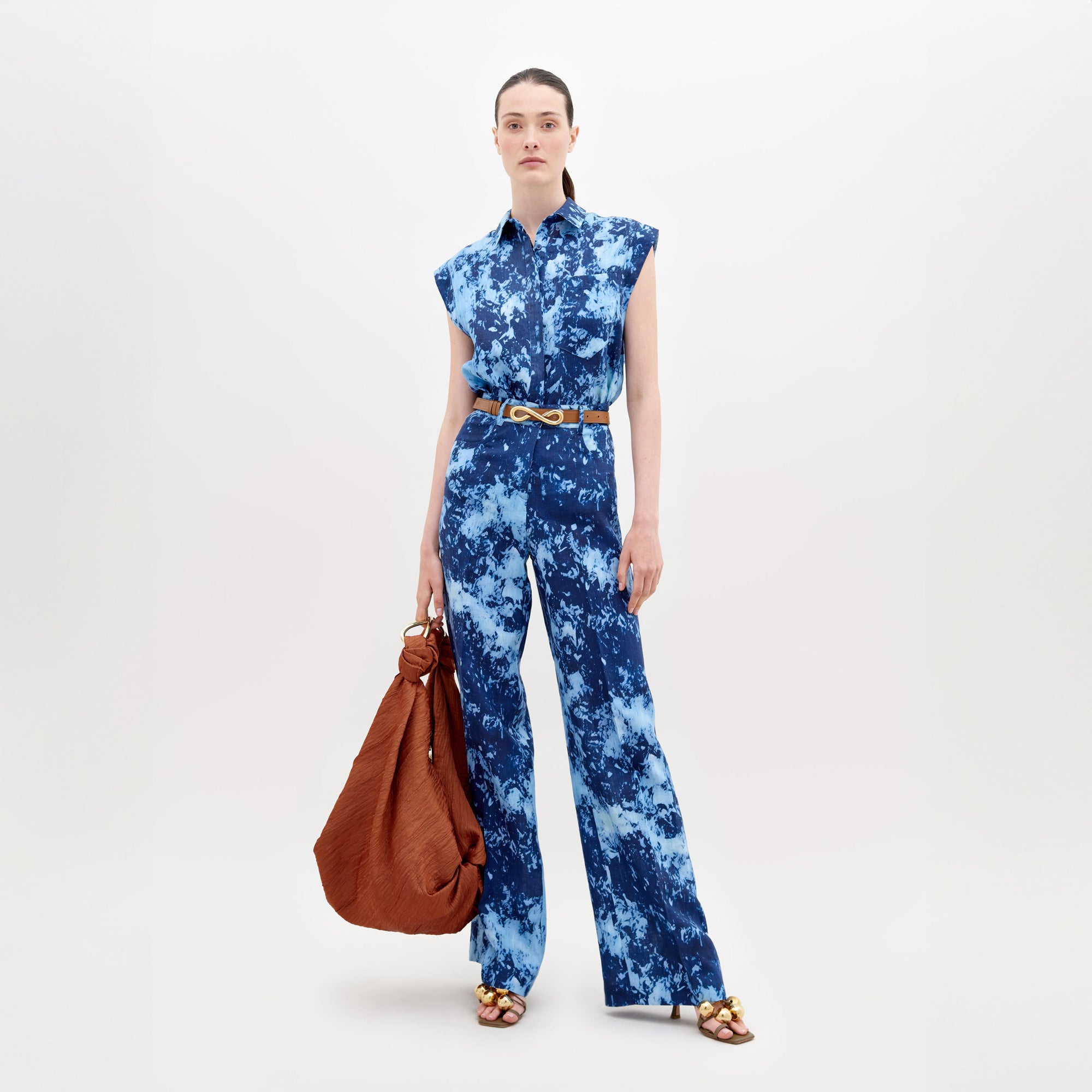 A person models the SVT DESIGNS Emine Pant, a blue sleeveless tie-dye jumpsuit with wide-leg trousers and a gold belt. They carry a large brown bag and wear open-toed sandals featuring gold accents, set against a simple light background.