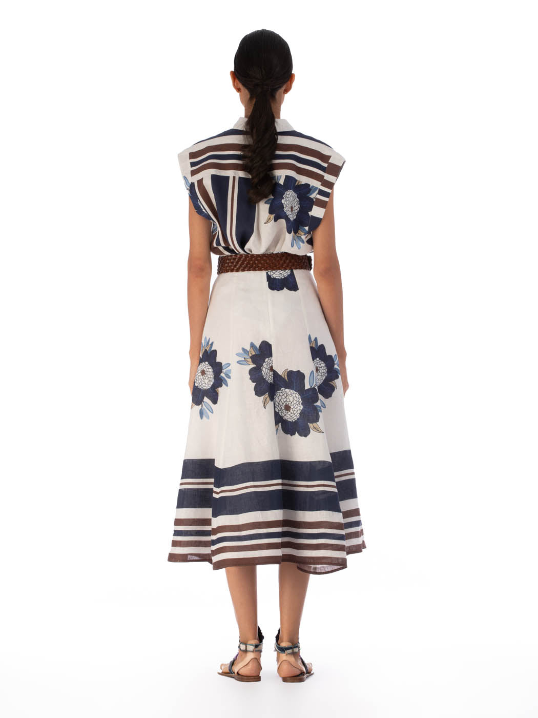 A person with long hair stands facing away, wearing the Erin Skirt from SVT DESIGNS, featuring blue floral patterns and brown stripes. The high-waisted midi skirt flows gracefully, paired effortlessly with flat sandals for a breezy look.