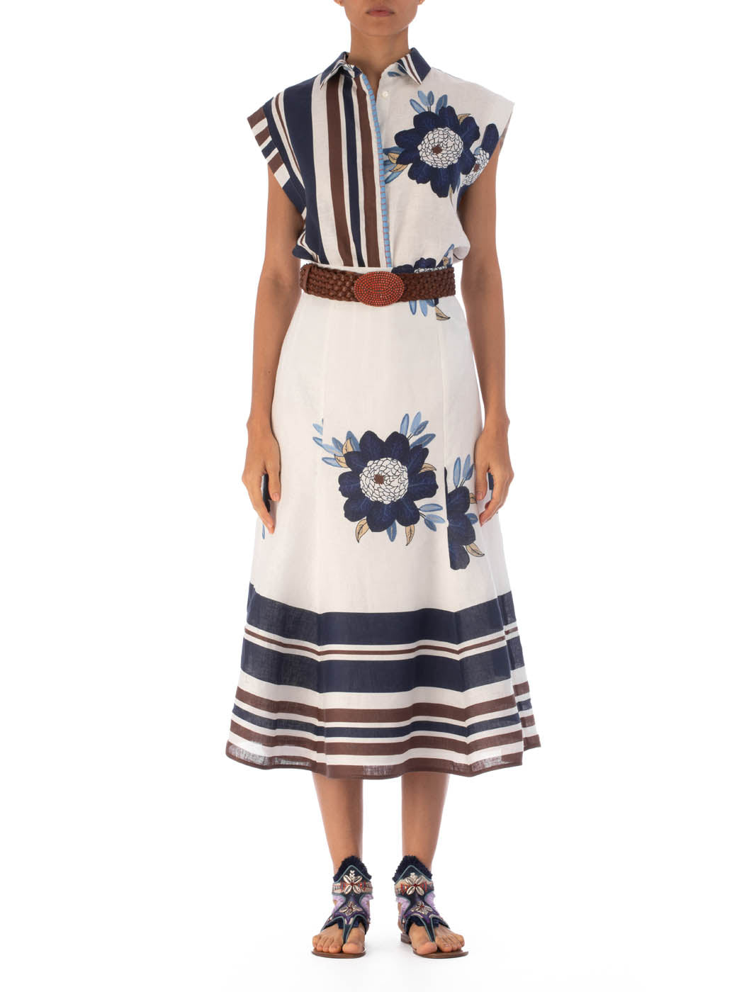 A person wearing the Erin Skirt by SVT DESIGNS, a sleeveless white dress with blue floral prints and vertical brown and blue stripes, exudes elegance. The 100% cotton outfit includes a brown belt and blue sandals against a plain white background.
