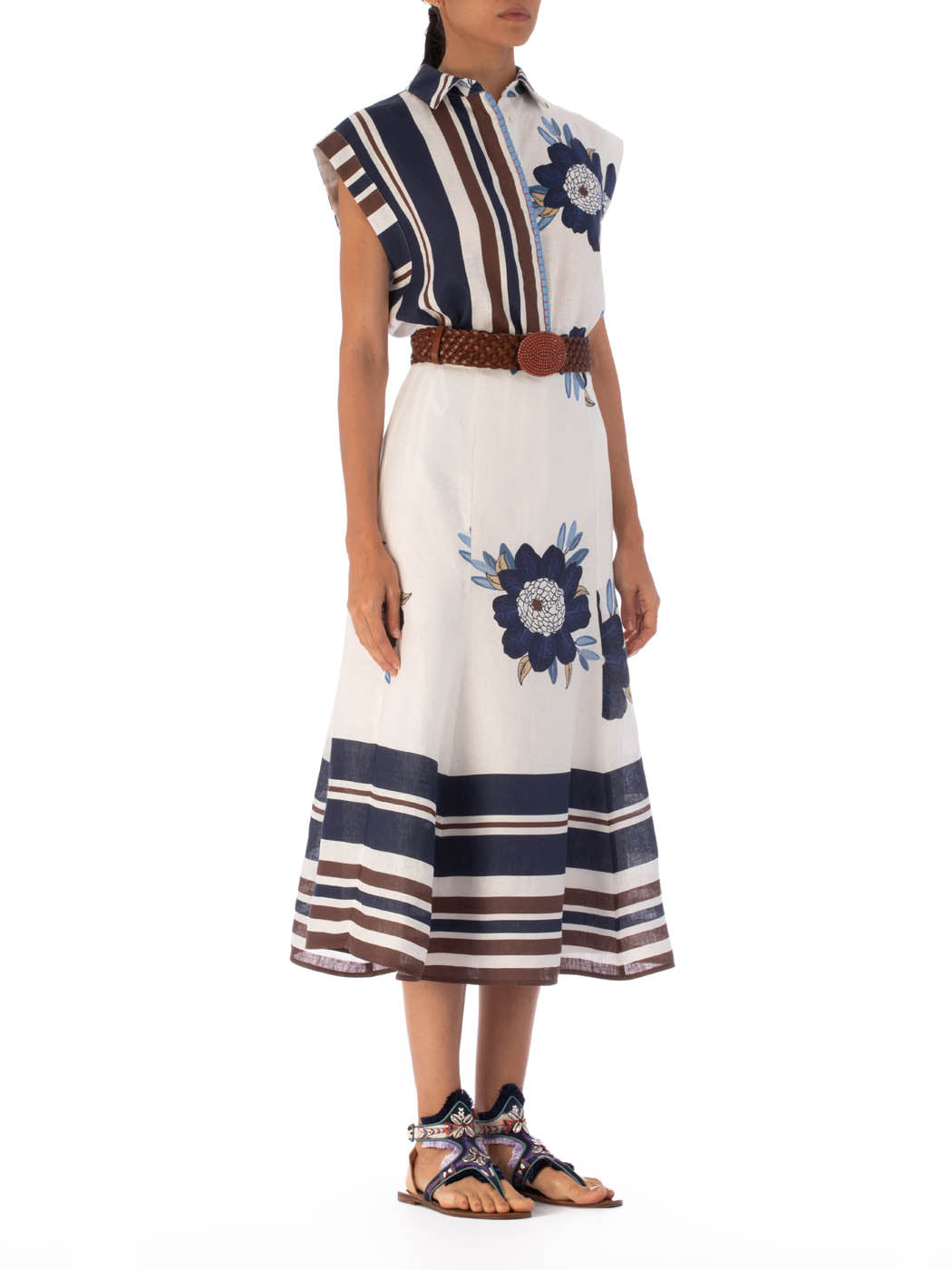 Someone is wearing the Erin Skirt by SVT DESIGNS, a sleeveless dress with blue and brown stripes featuring large floral prints, made from 100% cotton. A brown braided belt completes the high-waisted design, paired with open-toed sandals adorned with colorful straps on a plain white backdrop.