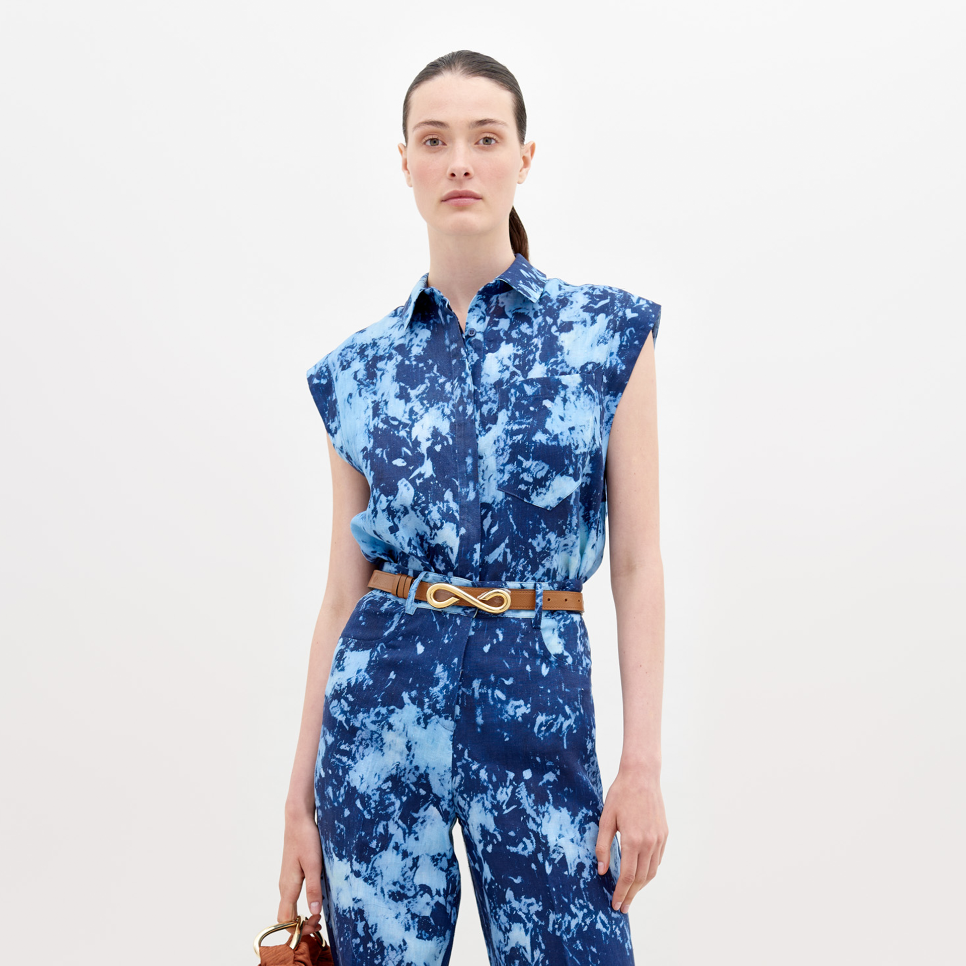 A woman stands against a white background, wearing the SVT DESIGNS Rina Blouse. It features Italian textiles in blue tie-dye, is sleeveless with two chest pockets and a tan belt. She holds a brown handbag and her hair is tied back.