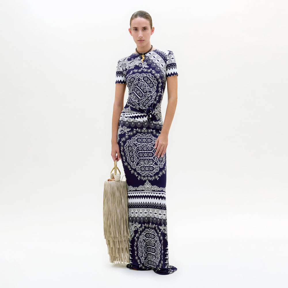 Woman wearing a long, patterned dress holding a woven bag on a white background