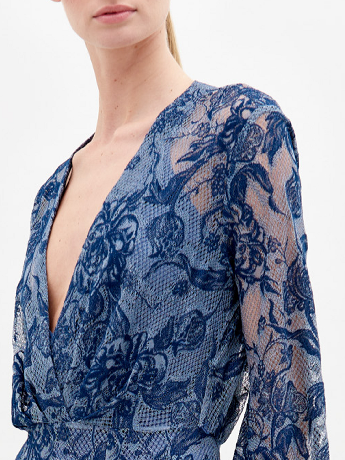 A person in a long, blue lace dress with a deep V-neckline and long sleeves stands against a plain white background, holding a black clutch—the Robin Dress Indigo Lace, available for pre-order, set to ship by November 15th, 2024.