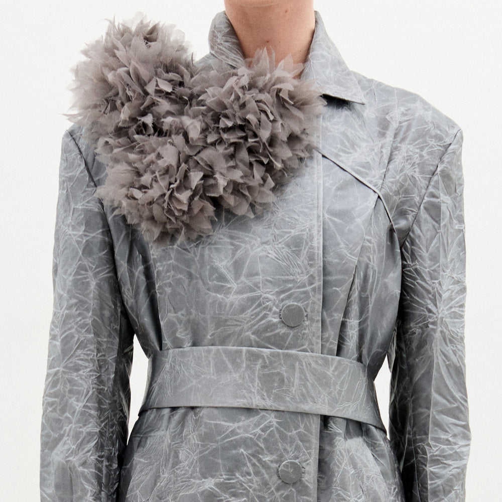 A person wears a crinkled, silver-gray Italian-fabric trench coat accented with SVT DESIGNS' Romy Collar Piece, a large textured gray floral embellishment on the left shoulder. The background is plain and light-colored.