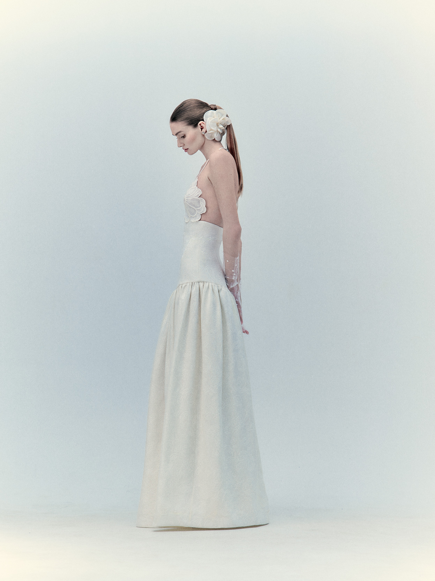 A woman in a Ruth Dress Pearl gown, elegant white with floral embellishments, wearing a decorative hairpiece, stands side profile against a light backdrop.