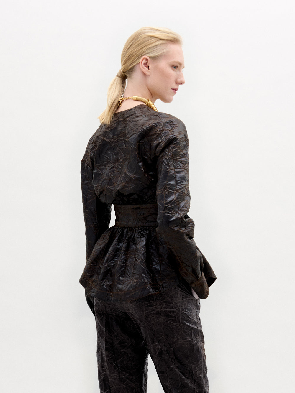 A blonde with a ponytail models the textured dark Saanvi Blouse and matching pants by SVT DESIGNS, featuring Italian fabric and a peplum waist. They're wearing a chunky necklace on a plain white backdrop, turned slightly away from the camera.