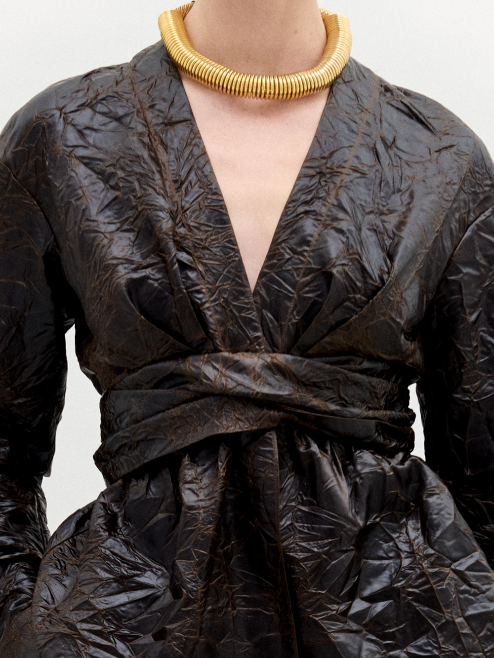 A person stands against a plain white background, wearing the Saanvi Blouse—a textured dark brown wrap top with a fitted waist—paired with matching pants and a chunky gold necklace.