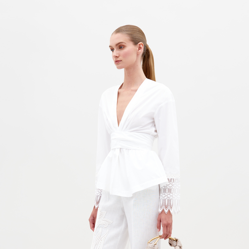 A woman stands against a plain background wearing the SVT DESIGNS Saanvi Blouse, featuring lace trim, paired with matching white pants. She holds a handbag with a gold handle and has her hair tied back in a sleek ponytail.