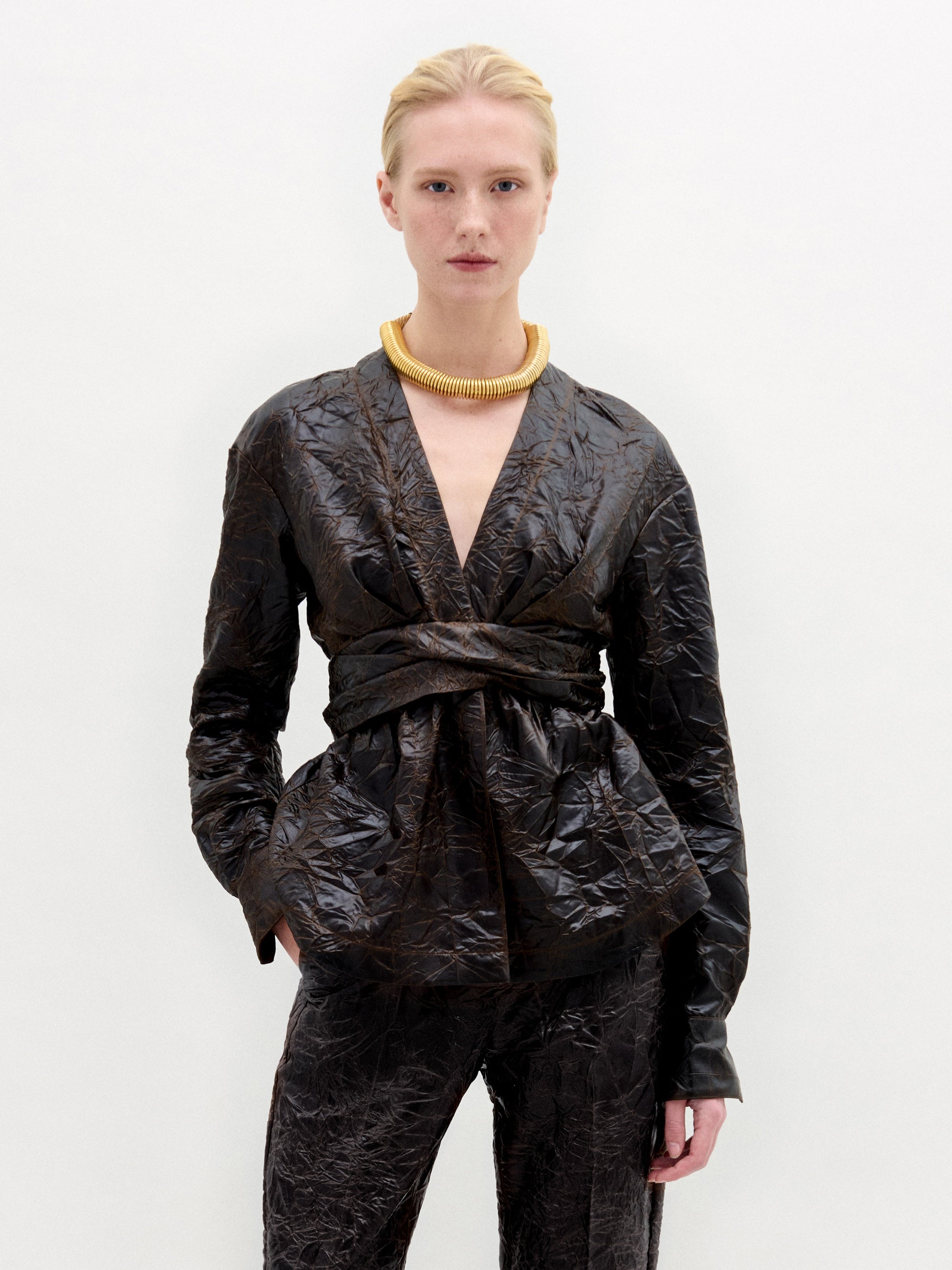 A person with light skin and blonde hair pulled back wears the Saanvi Blouse by SVT DESIGNS—a shiny, dark brown textured blouse with a fitted waist—paired with matching pants and a chunky gold necklace, against a plain white background.