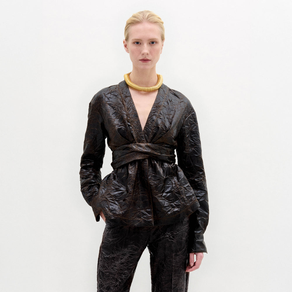 A person stands against a plain white background, wearing the Saanvi Blouse—a textured dark brown wrap top with a fitted waist—paired with matching pants and a chunky gold necklace.