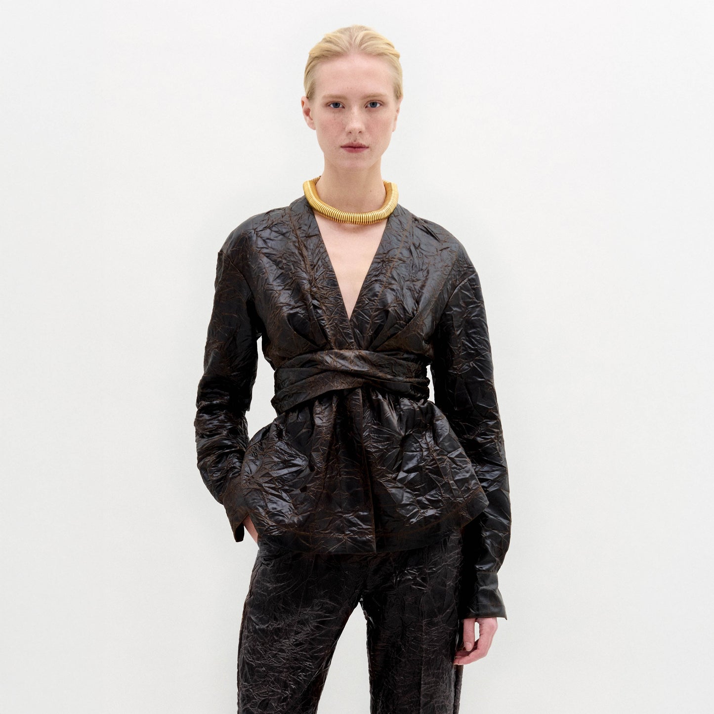 A person stands against a plain white background, wearing the Saanvi Blouse—a textured dark brown wrap top with a fitted waist—paired with matching pants and a chunky gold necklace.