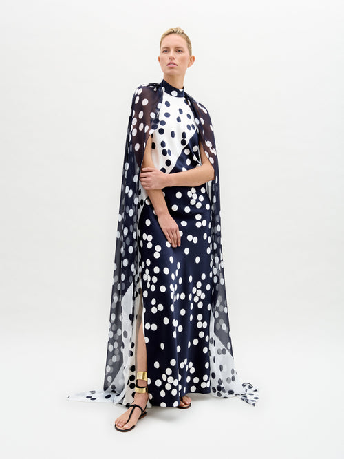 Against a plain white background, a person wears the Sabina Dress by SVT DESIGNS, featuring a navy and white polka-dotted fitted silk maxi dress with matching sheer cape, complemented by their short blonde hair and black sandals with gold details.