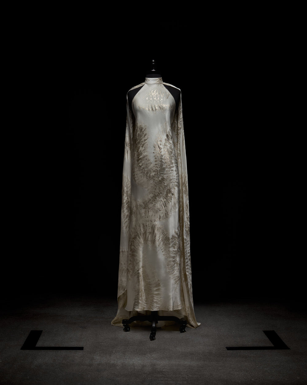 The Sabina Dress by SVT DESIGNS is showcased on a black mannequin in dim lighting. Its cream color features intricate, fern-like silver designs, and the dark floor accentuates its elegance.
