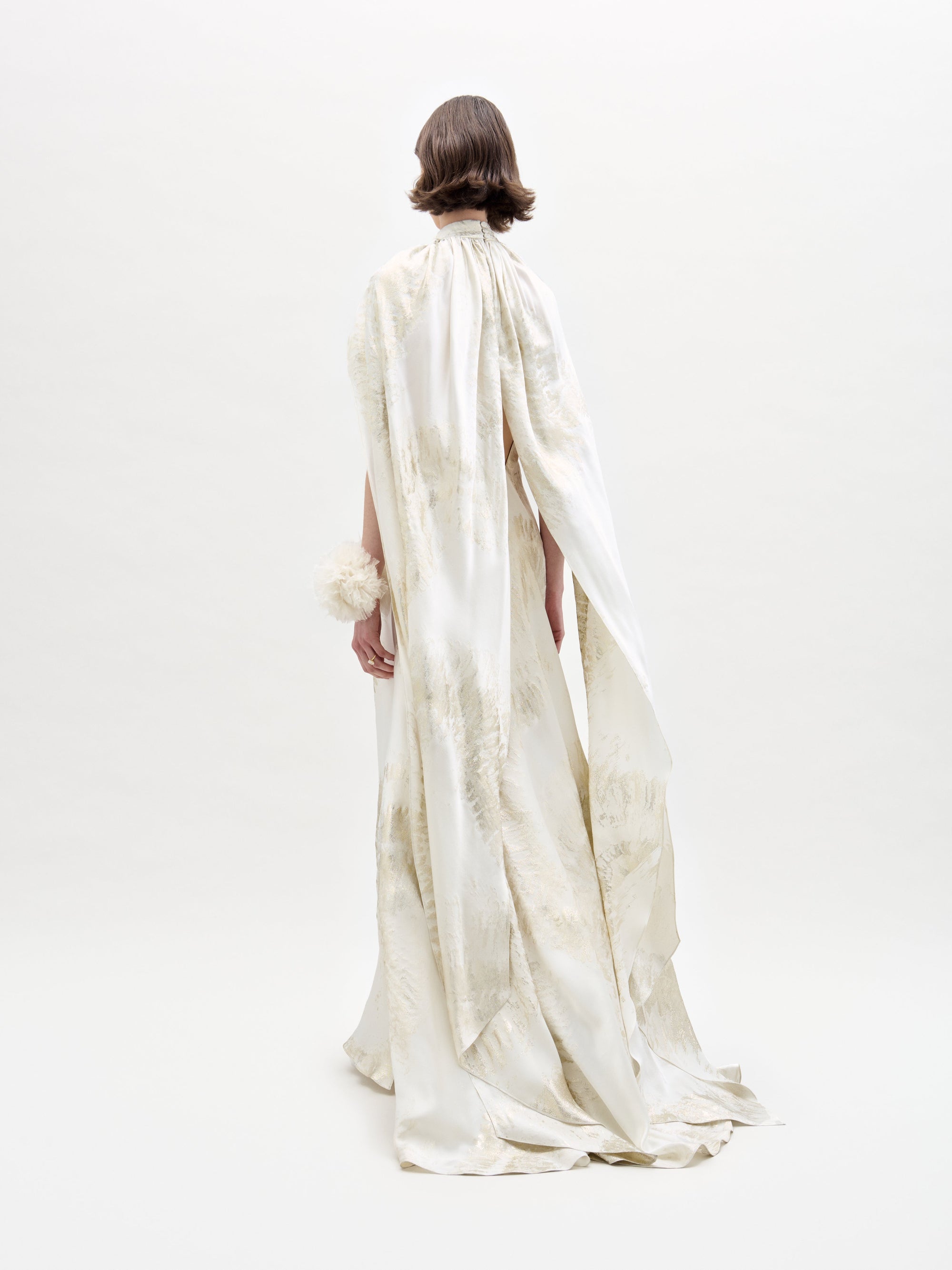 On a light background, a person faces away, wearing the Sabina Dress by SVT DESIGNS. This long, flowing white dress features textured patterns with a high collar and a cape-like back extension. Their hair is short and wavy.