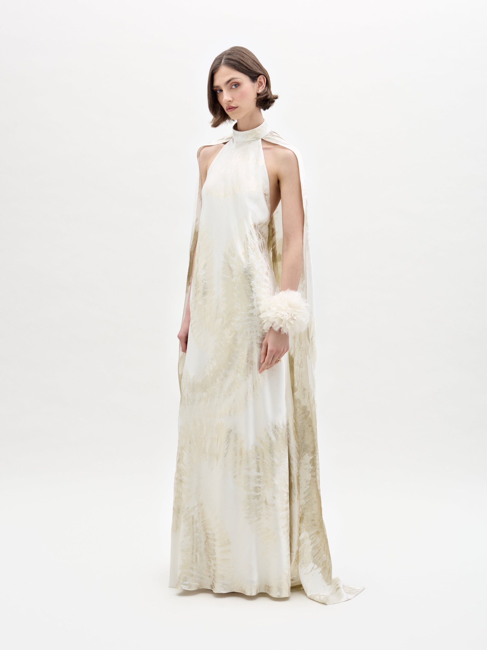 A woman wears the Sabina Dress by SVT DESIGNS, a long, flowing white dress with soft golden patterns, featuring a high neck and cape-like sleeves. She accessorizes with a fluffy white wrist piece against a plain white background.