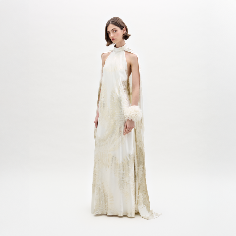 A woman with short brown hair and a neutral expression wears the Sabina Dress by SVT DESIGNS, featuring light beige patterns, a draped neck detail, and a fluffy white cuff, standing against a plain white background.