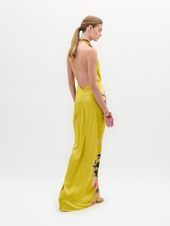 A person with long hair tied back stands in profile, wearing the Sabrina Dress by SVT DESIGNS. This yellow midi dress features floral patterns at the hem and is crafted from luxurious Italian fabric, standing out against a plain white background.