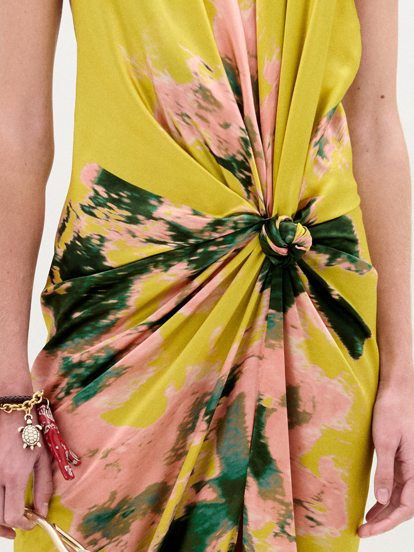 A person wears the Sabrina Dress by SVT DESIGNS, featuring a knot detail and crafted from luxurious Italian fabric. The vibrant abstract print in yellow, pink, and green stands out. In their left hand is an object with a pink strap, complemented by a charm bracelet on the wrist.