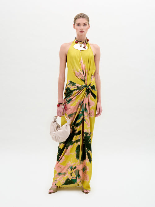 A person wearing the SVT DESIGNS Sabrina Dress, a long sleeveless yellow midi with an abstract pink and green floral pattern and central knot detail, stands against a plain background. They carry a cream clutch, wear sandals, have hair tied back, and are adorned with a necklace and bracelet for understated elegance.