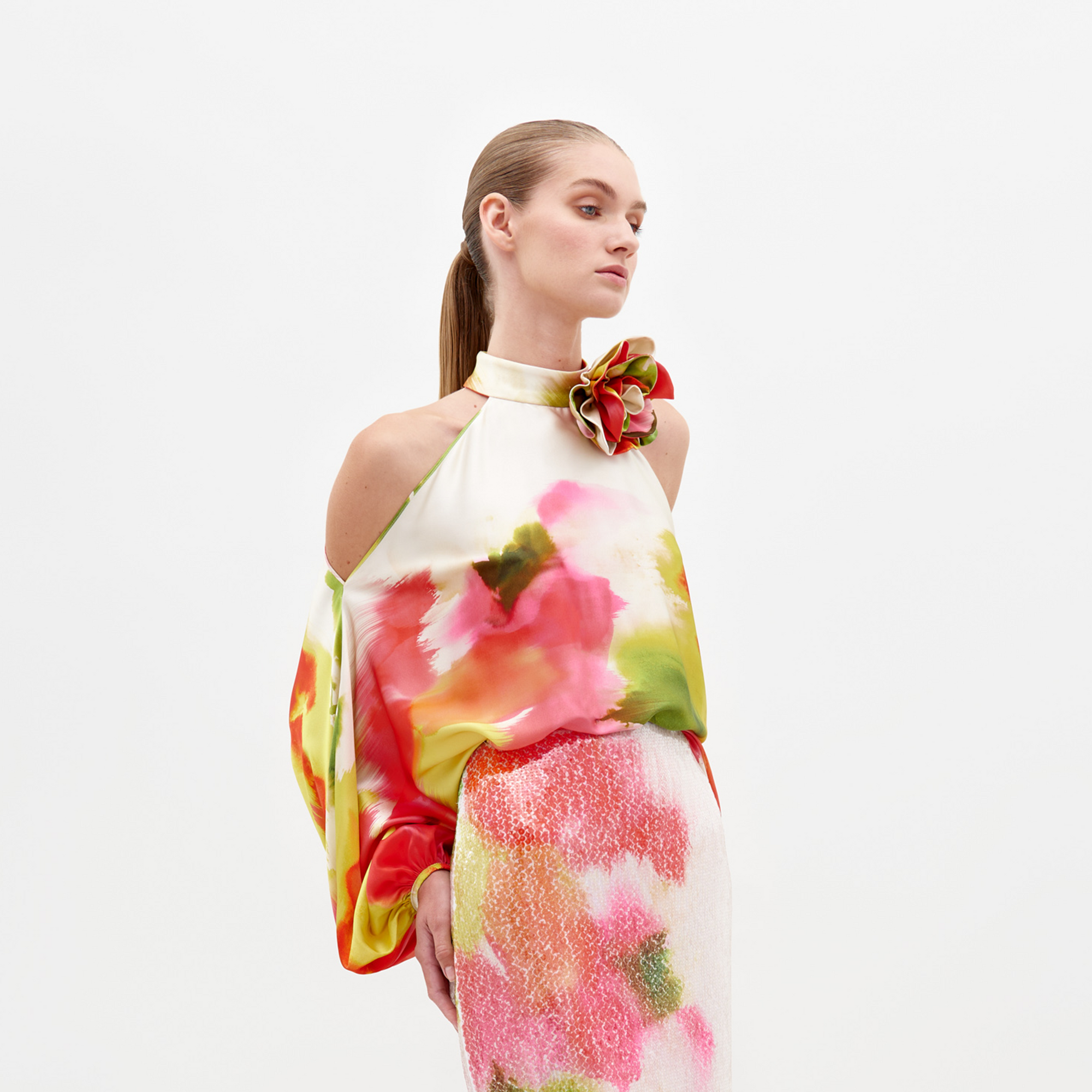 A person stands against a white background wearing an outfit with Italian textiles from SVT DESIGNS. The sleeveless Safira Blouse, featuring a halter neck and large floral appliqué on the shoulder, pairs seamlessly with the vibrant pink and green skirt.