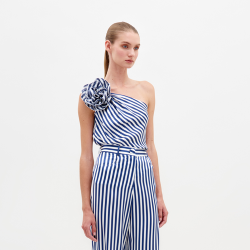 A person stands against a white background wearing the Sebastiana Blouse by SVT DESIGNS, an asymmetrical blue and white striped one-shoulder jumpsuit with a chic floral appliqué on the shoulder, enhanced by a matching belt for added sophistication.