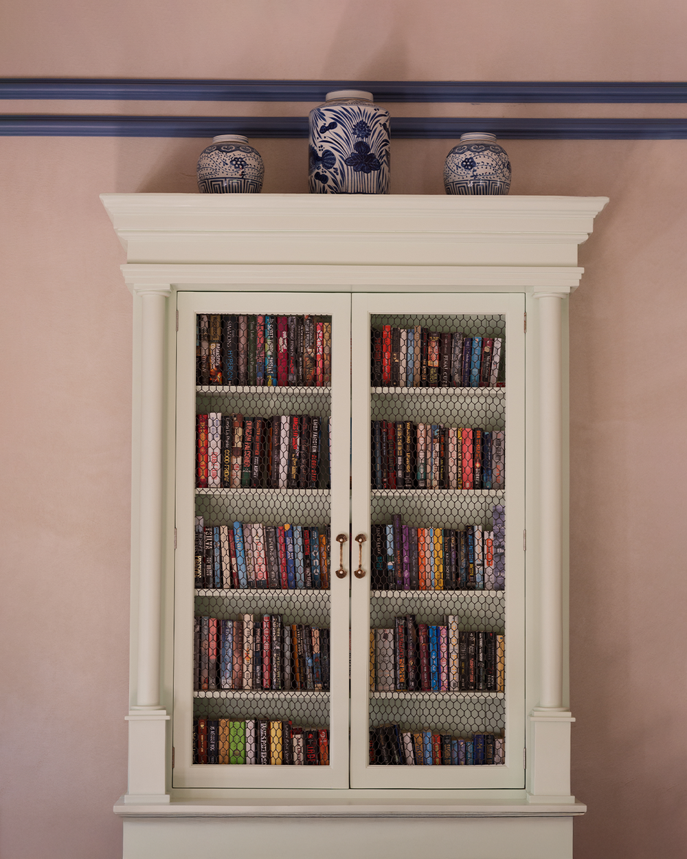 White bookshelf with glass doors displaying books against a pink wall.