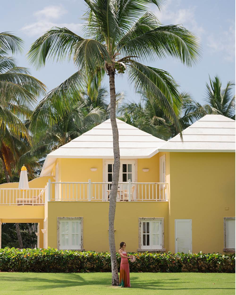 Yellow villa with a palm tree in front, surrounded by greenery and blue sky.