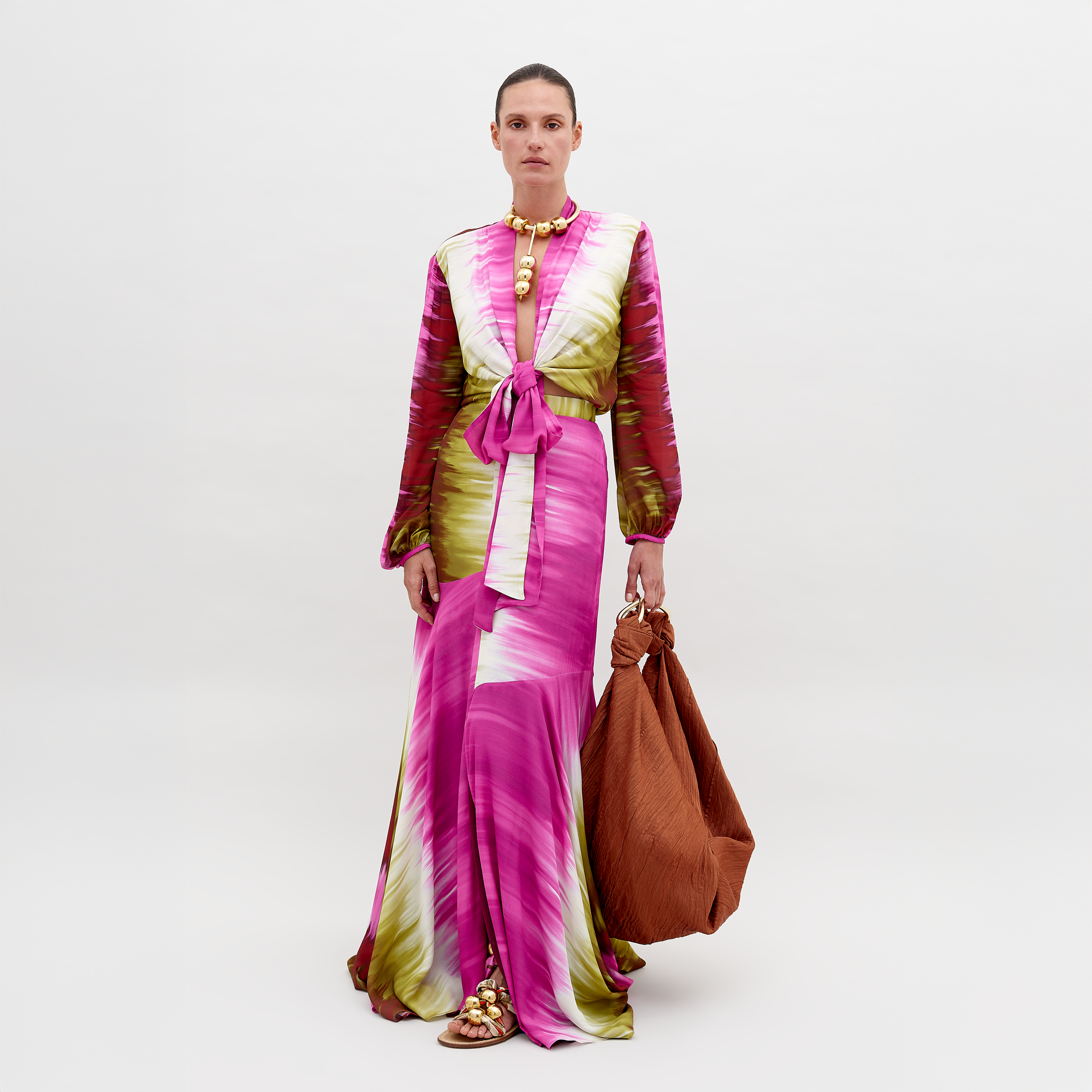 Against a white background, an individual wears the Shane Skirt by SVT DESIGNS, characterized by vibrant shades of pink, white, and green. A tied belt adorns the waist with gold embellishments at the neck and hem. The flared hem gracefully skims the floor as they carry a large textured brown bag.