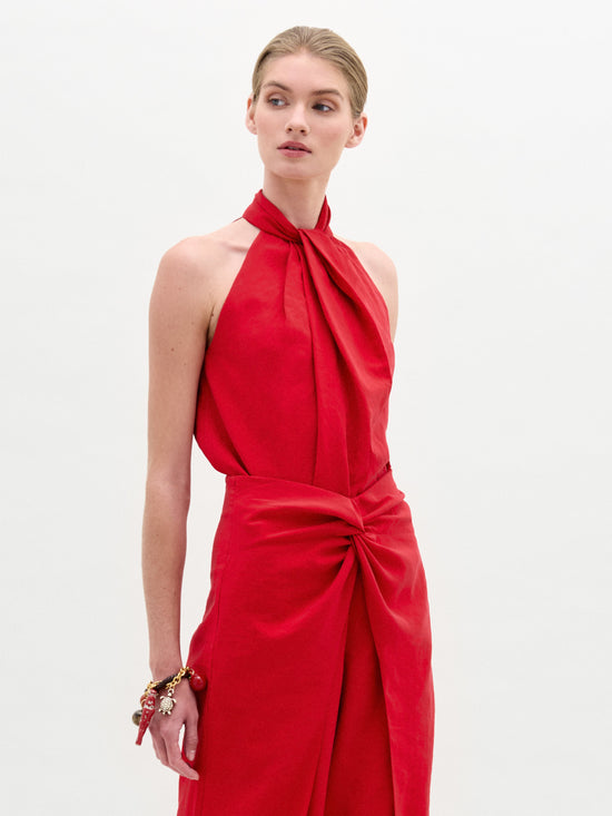 A person in a sleeveless red SVT DESIGNS dress made from Italian fabric stands against a plain background. Featuring a halter neck and waist twist detail, the Shela Blouse complements their bracelet-adorned left wrist as they gaze slightly to the side.