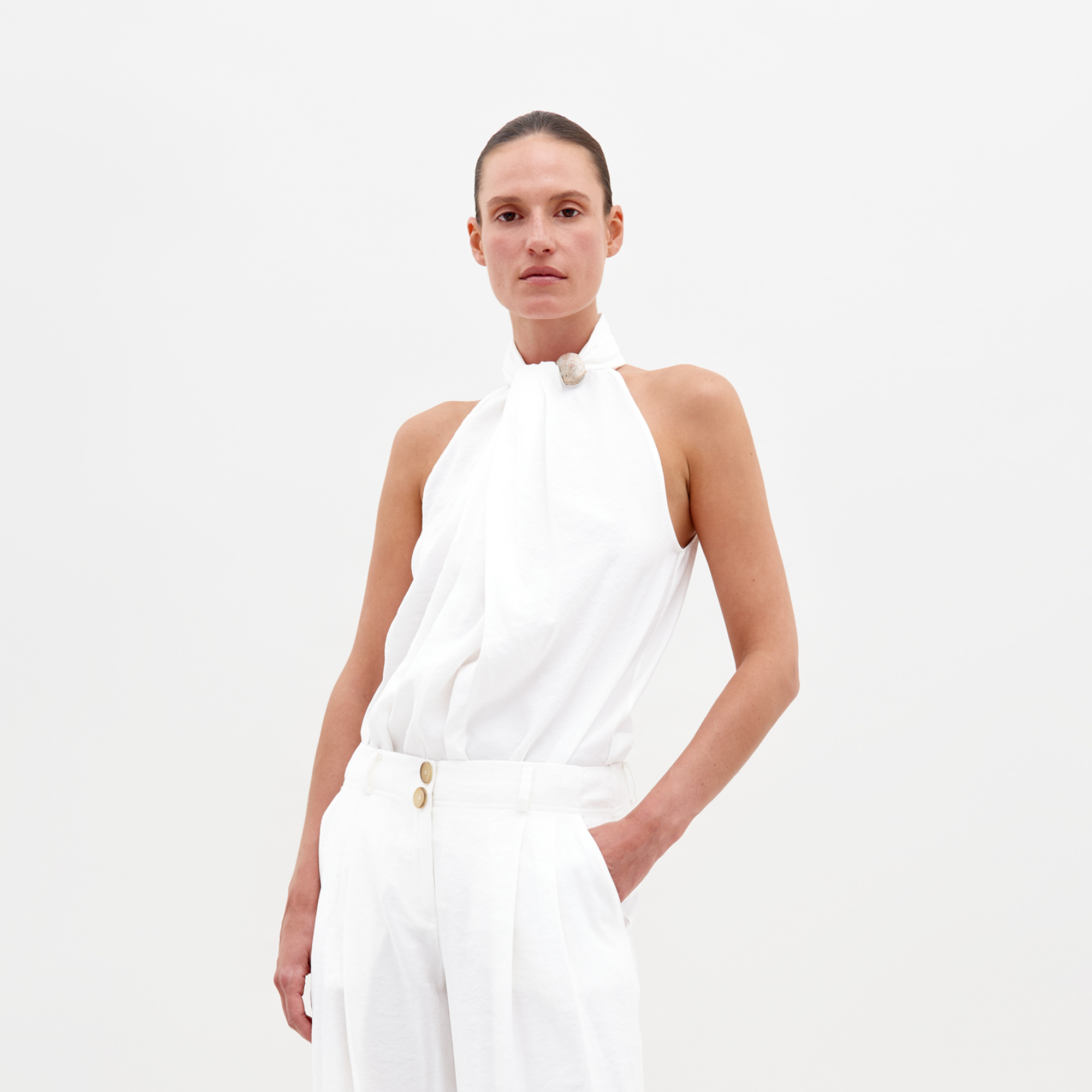 A person with tied-back hair is wearing the Shela Blouse by SVT DESIGNS and white pants. This white halter-neck blouse features a draped design and an embellished neckline, elegantly highlighted against a plain white backdrop showcasing Italian textiles.
