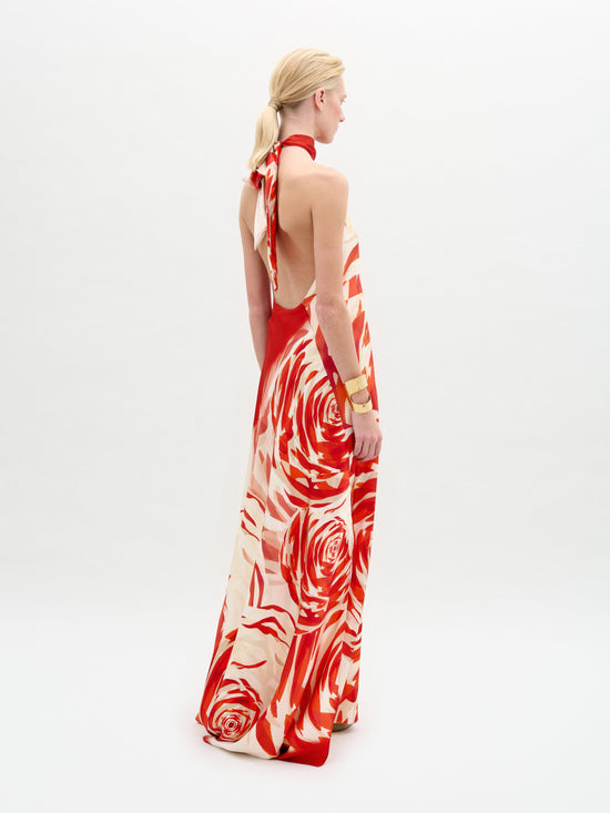 A blonde woman faces slightly right, wearing the sleeveless, backless Sherry Dress by SVT DESIGNS. The silk maxi dress has a red and cream floral pattern with a halter neckline. She sports a bracelet on her left arm against a plain white background.