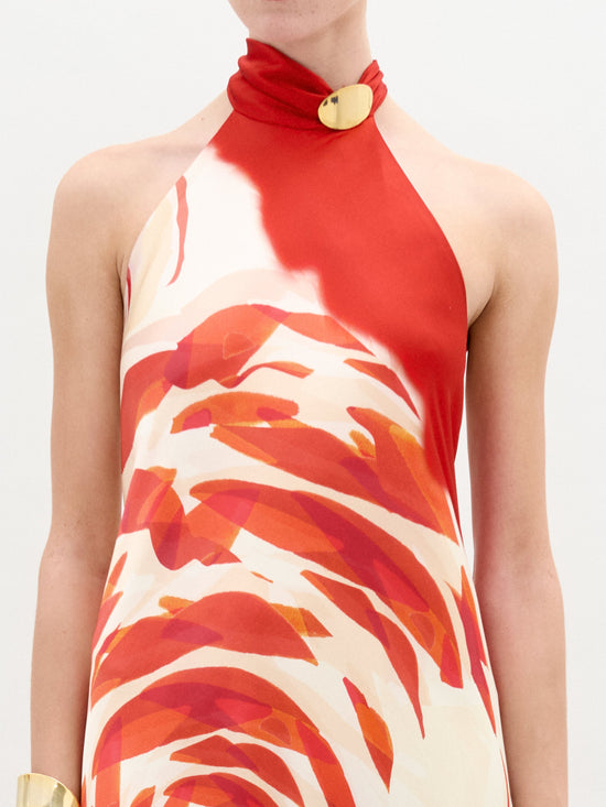 A person is elegantly wearing the Sherry Dress by SVT DESIGNS, a sleeveless silk maxi featuring bold red and orange abstract patterns. The unique dual-buttoned collar and halter neckline crafted from fine Italian fabric perfectly complement their bare arms against a plain white background.