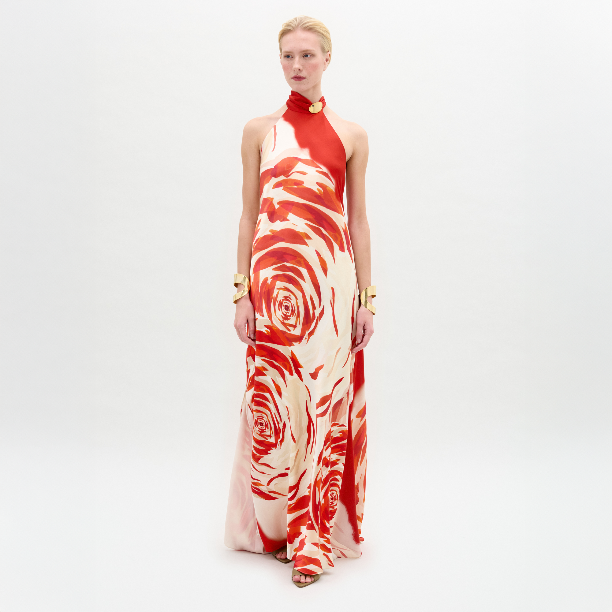 A person with pale skin and blonde hair pulled back wears the SVT DESIGNS Sherry Dress, a sleeveless red and white silk maxi with bold rose pattern and halter neckline, styled with gold bracelets, against a plain white background.