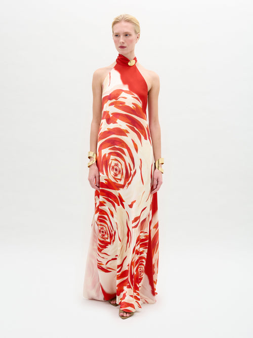 A person models the elegant SVT DESIGNS Sherry Dress, a sleeveless, high-neck gown made from luxurious Italian fabric with a striking red and cream abstract rose pattern. They have short blonde hair, accessorized with chunky gold bracelets on a plain white background.