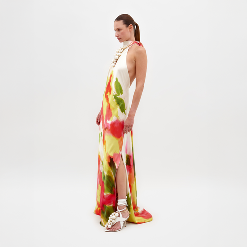 A person stands against a plain white background wearing the Sherry Dress by SVT DESIGNS. It's a sleeveless, colorful maxi silk dress with an abstract print in red, green, and yellow patterns featuring a side slit. They pair it with white high-heeled sandals adorned with large decorative beads.