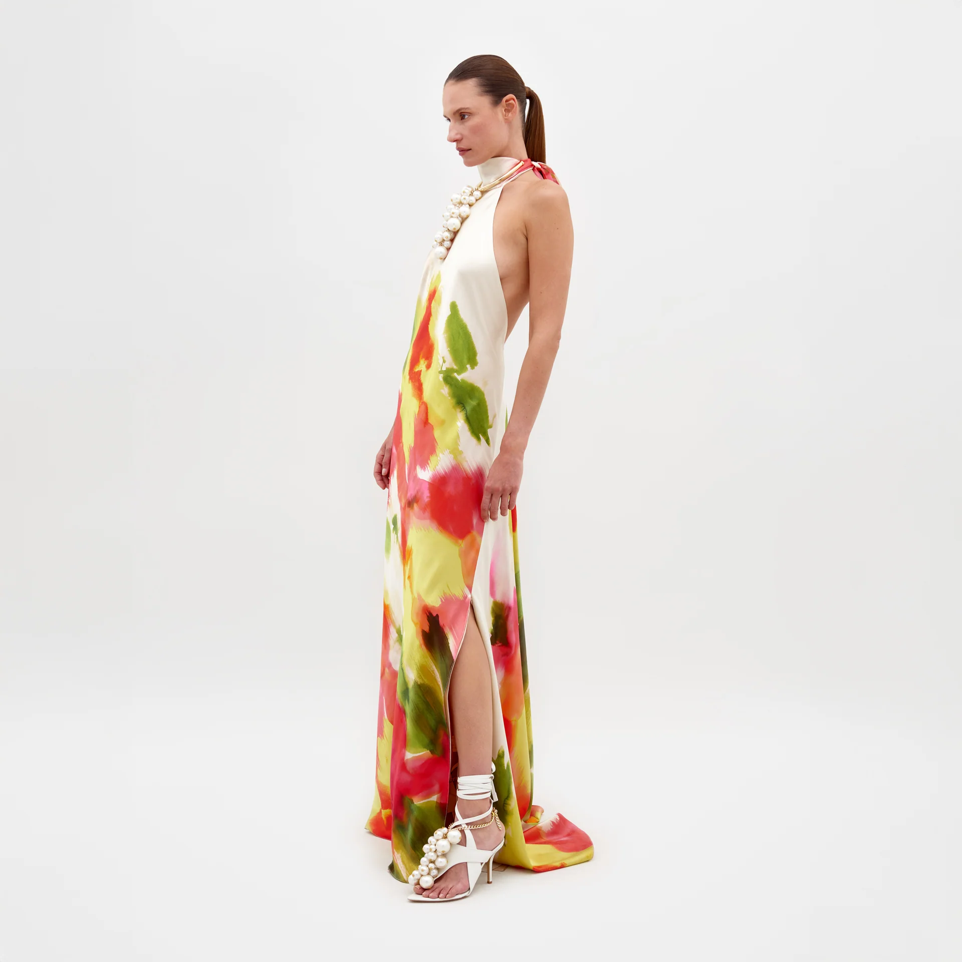 A woman in a white room wears the Sherry Dress by SVT DESIGNS, a vibrant silk maxi with abstract red, green, and yellow prints. She pairs it with slicked-back hair, white high-heeled sandals, and a chunky pearl necklace. Made in Colombia.