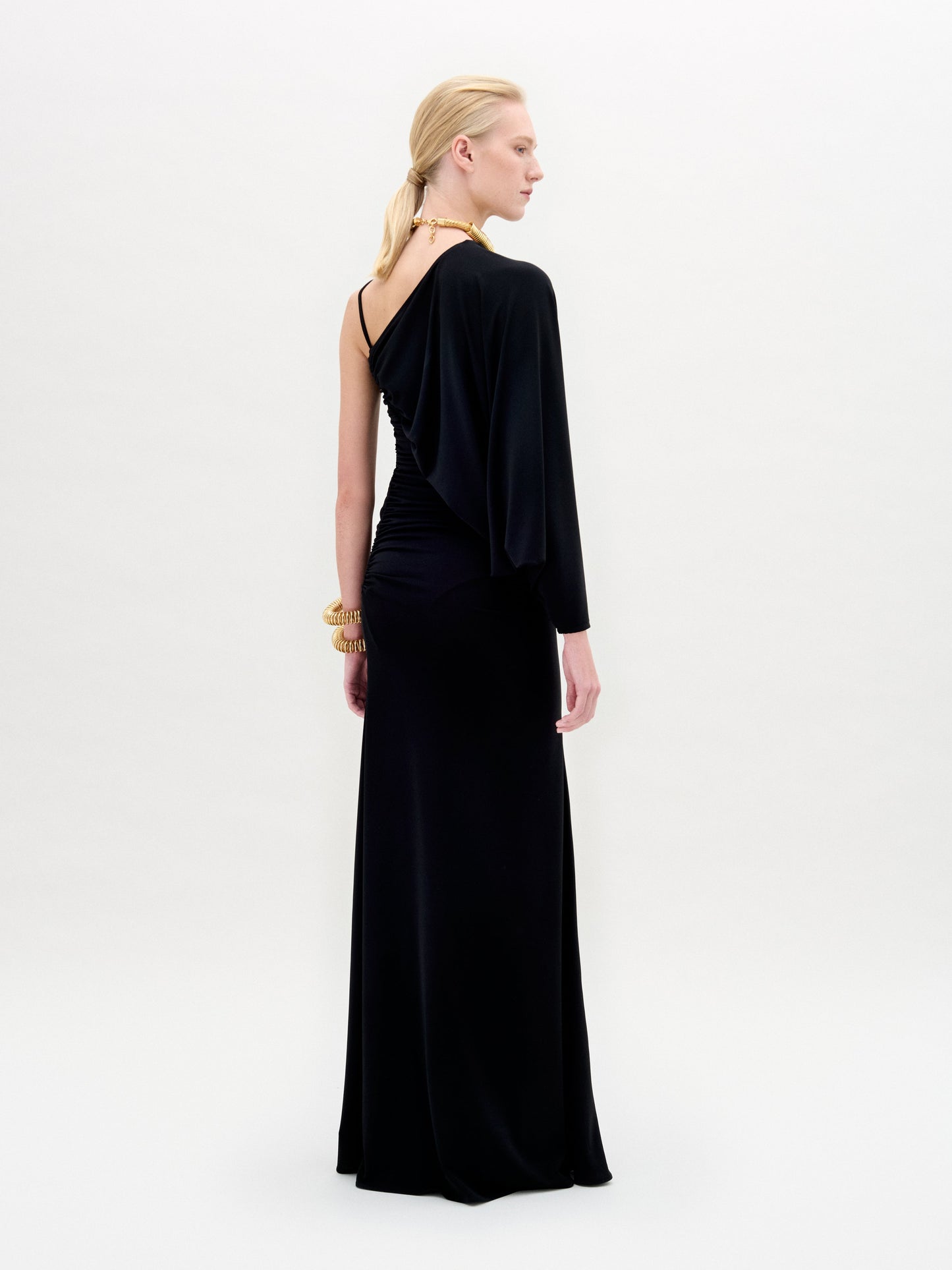 Against a plain background, a person showcases the Shiri Dress, a long black asymmetrical column silhouette crafted from Italian fabric, paired with gold accessories.