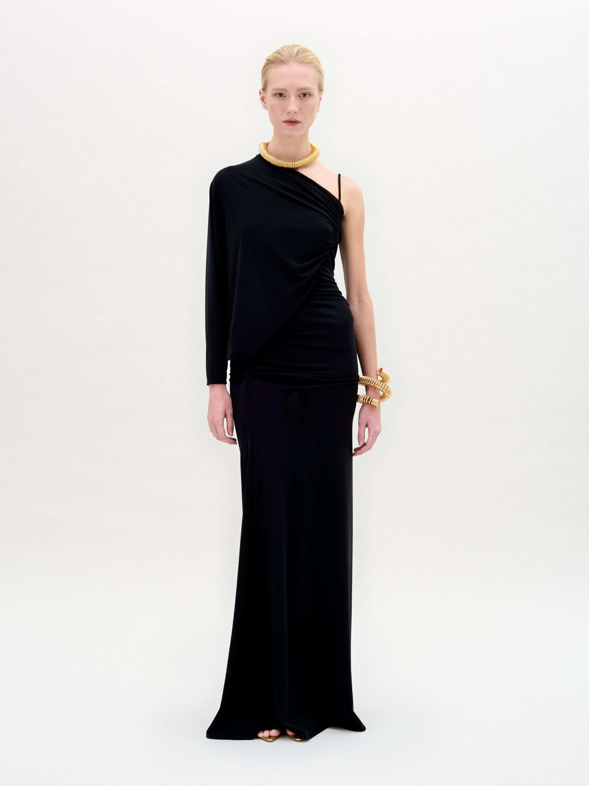 A woman stands against a white background wearing the SVT DESIGNS Shiri Dress, a black one-shoulder maxi with a column silhouette, styled with gold jewelry. She faces forward with a neutral expression.