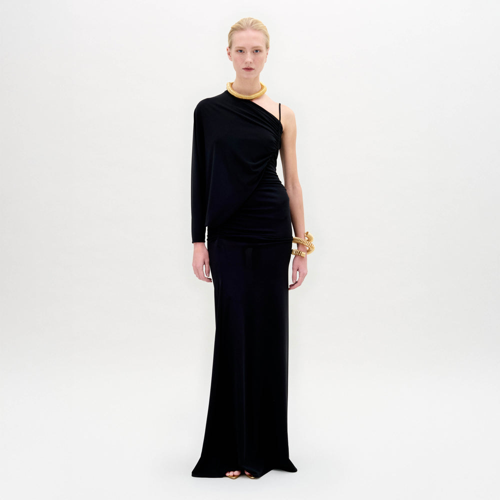 A person poses against a white background wearing the SVT DESIGNS Shiri Dress, a long black one-shoulder maxi with a column silhouette. They accessorize with gold jewelry and have their hair pulled back.