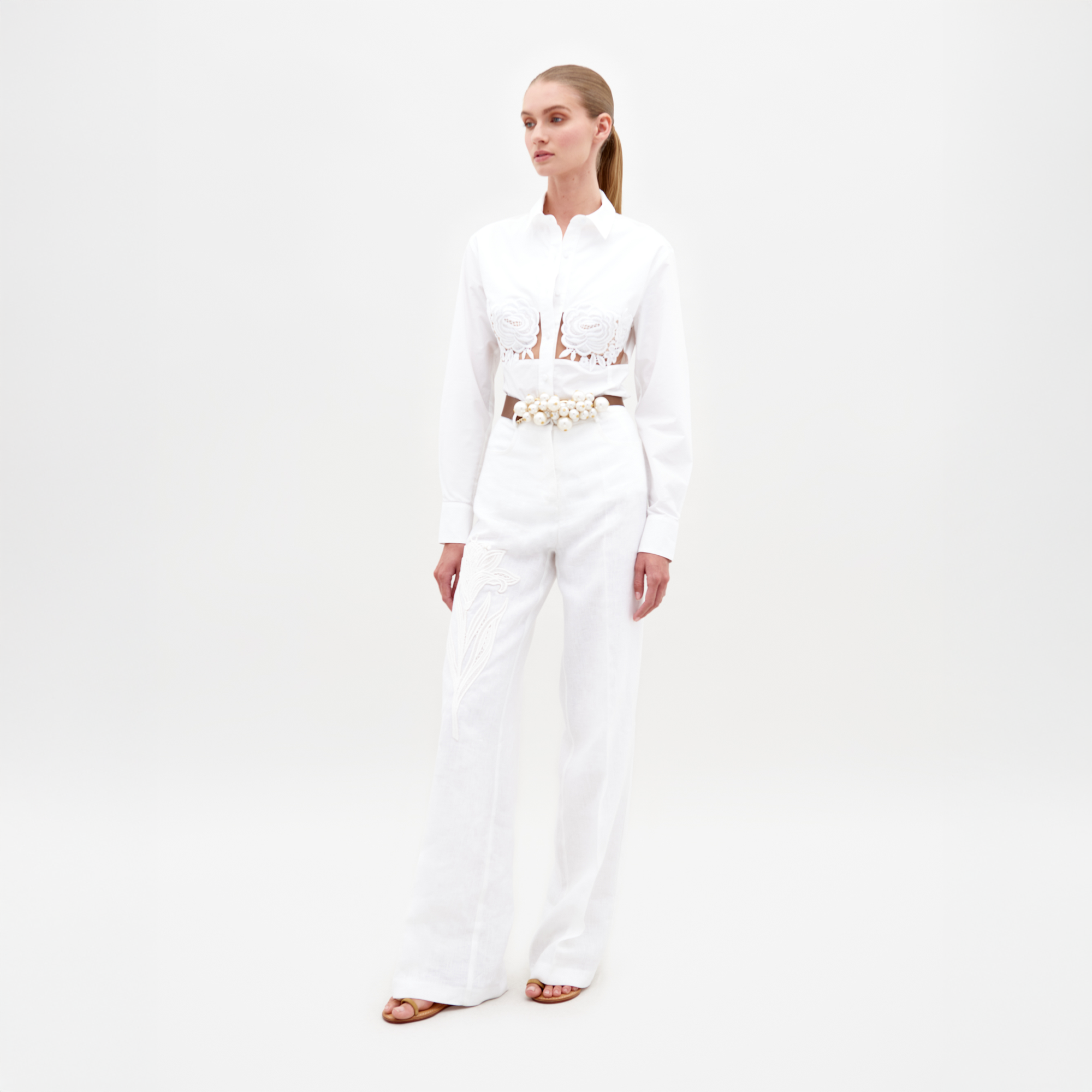 A woman stands against a white background wearing the Trilce Pant—high-waisted white linen pants with embroidery—paired with a white button-up shirt with cut-out details, a shell-embellished belt, and brown open-toe sandals.