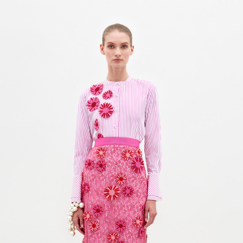 A woman stands against a plain white background, wearing the pink and white striped Silvina Blouse by SVT DESIGNS and a pink skirt with 3D floral appliqué. She accessorizes with a pearl bracelet and looks directly at the camera.